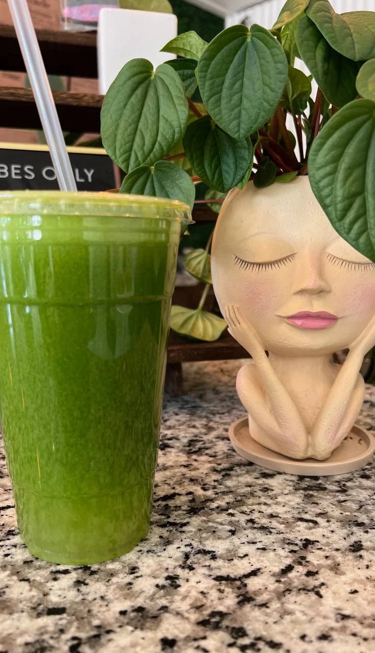 Green Raw South juice beside a leafy plant in a face-shaped planter